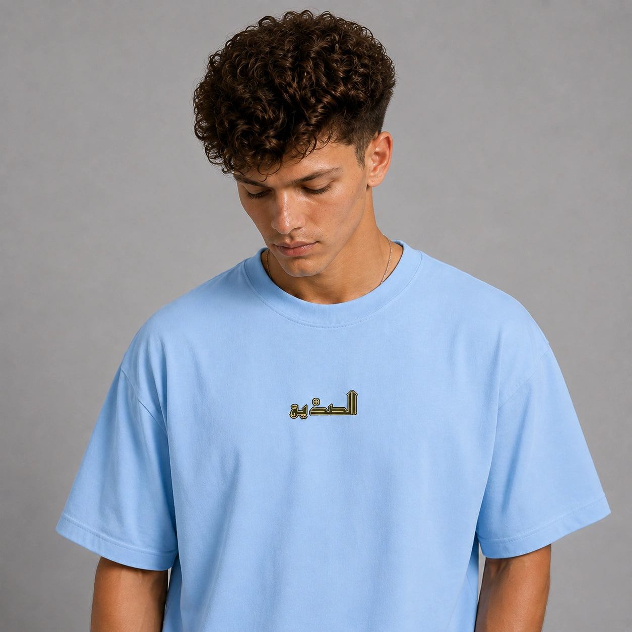 A man wearing a light blue crewneck t-shirt with gold Arabic calligraphy on the chest, photographed against a neutral gray background.