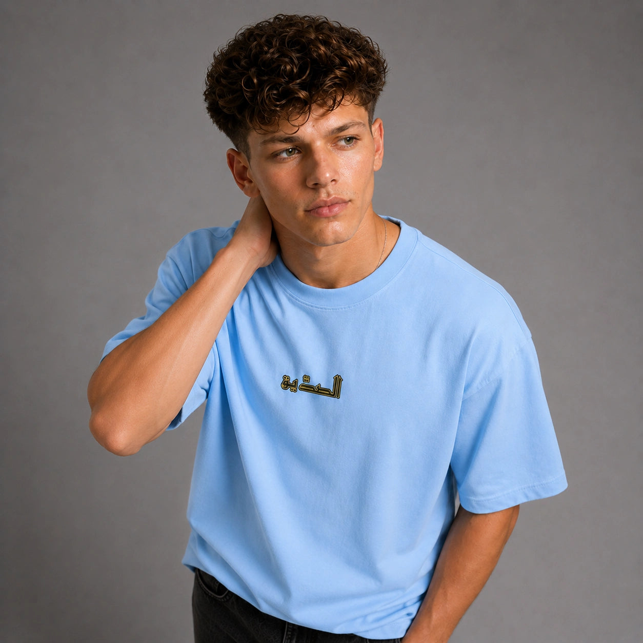 A young man with curly brown hair wearing a light blue crewneck t-shirt with black Arabic calligraphy on the chest, posing against a solid gray studio background.