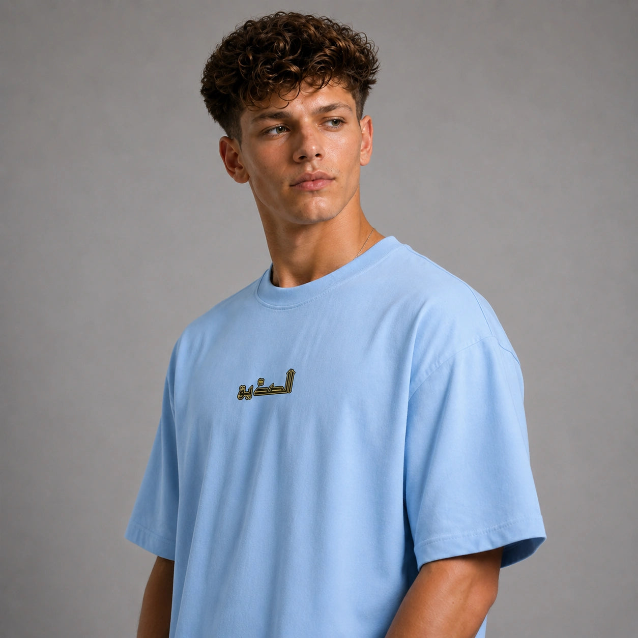 A young man with curly brown hair wearing an oversized light blue t-shirt with gold Arabic calligraphy on the chest, photographed against a neutral gray studio background.