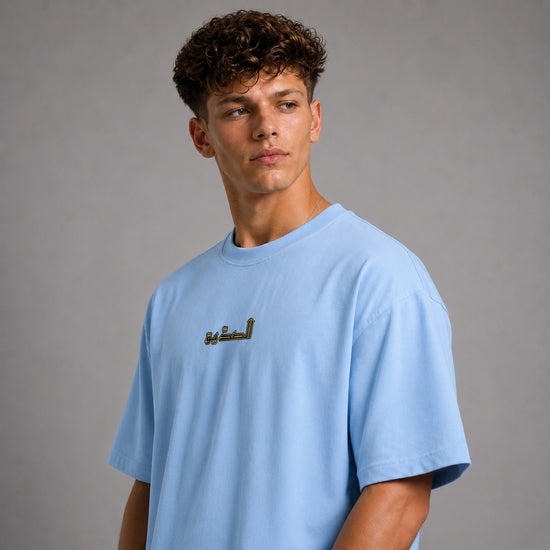 A young man with curly brown hair wearing an oversized light blue t-shirt with gold Arabic calligraphy on the chest, photographed against a neutral gray studio background.