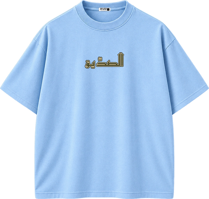 A light blue short-sleeve crewneck t-shirt with Arabic text printed in gold and black on the chest, displayed on a solid white background.