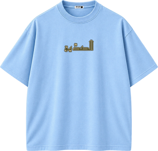A light blue short-sleeve crewneck t-shirt with Arabic text printed in gold and black on the chest, displayed on a solid white background.