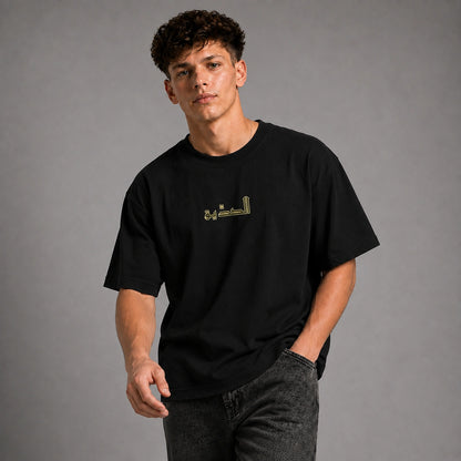 A young man wearing a black crewneck t-shirt with gold Arabic calligraphy on the chest, paired with dark gray jeans, standing against a solid gray studio background.