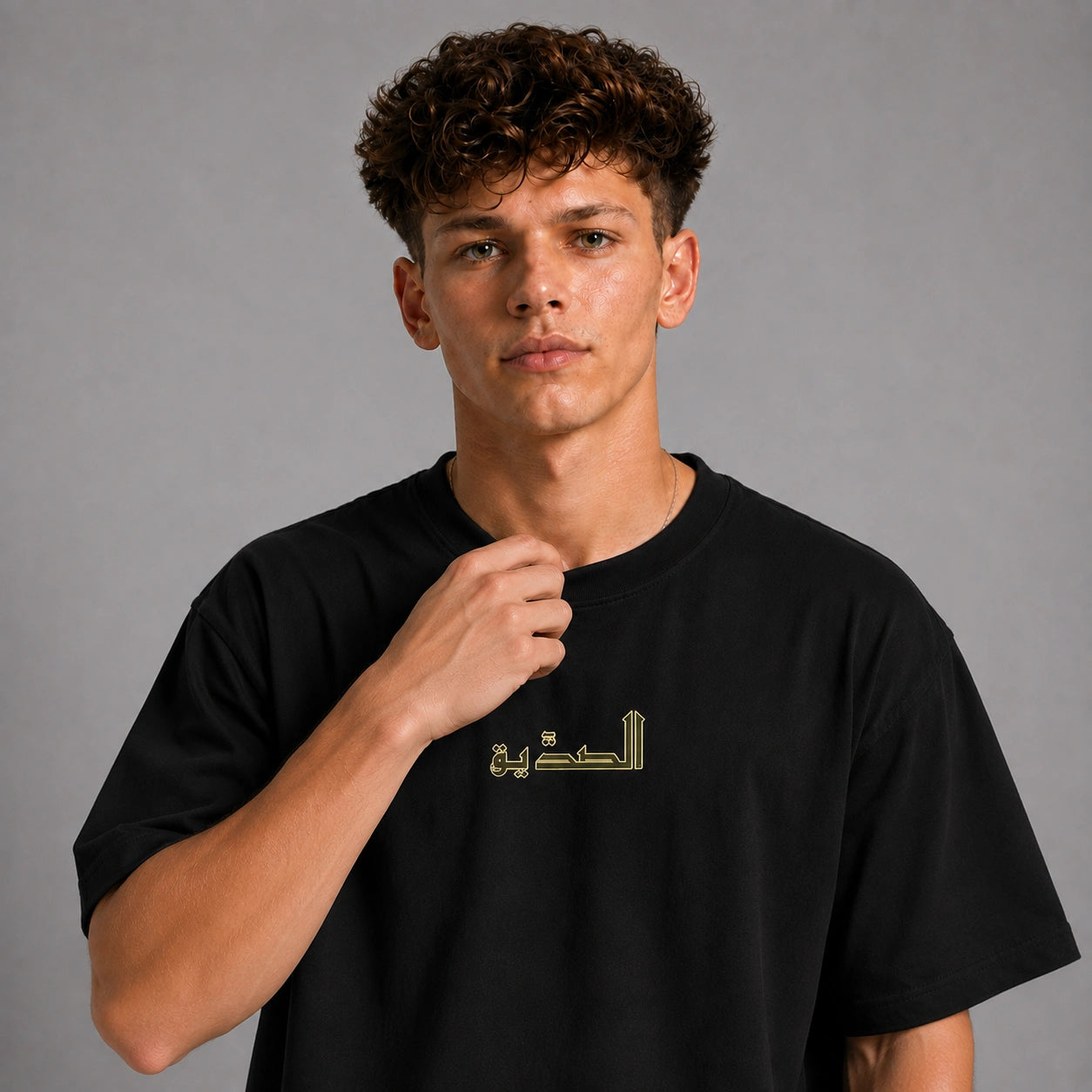 A young man with curly brown hair wearing a black short-sleeve t-shirt with gold Arabic calligraphy on the chest, posed against a neutral gray studio background.