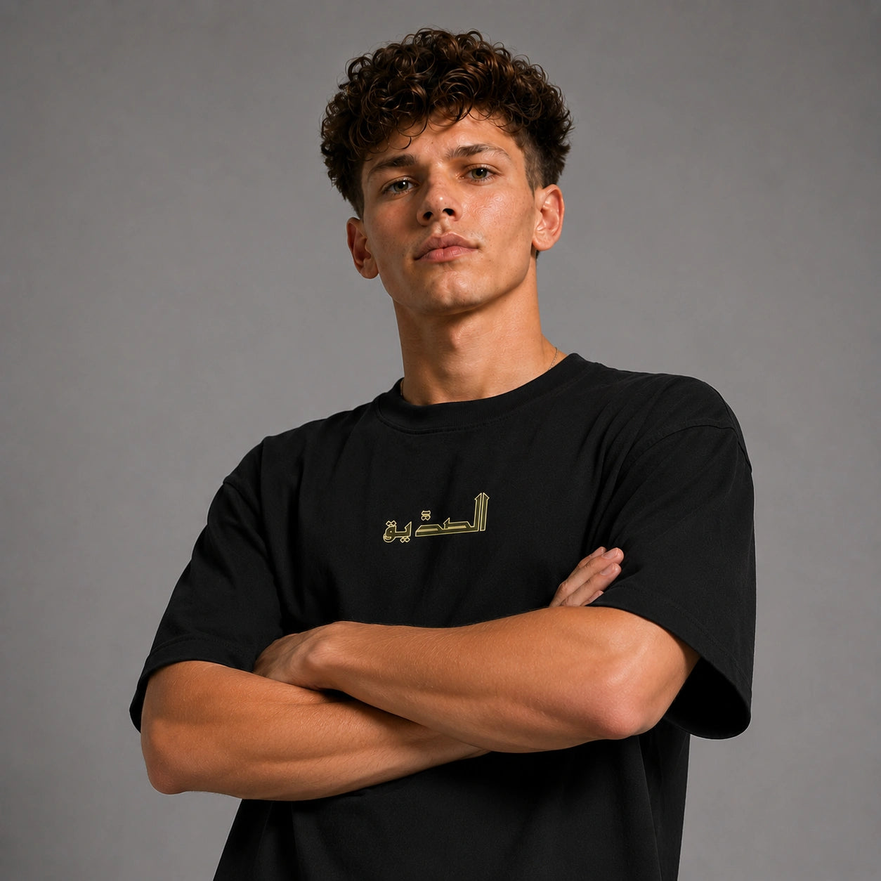 A young man with curly brown hair wearing a black crewneck t-shirt with gold Arabic calligraphy on the chest, posed with arms crossed against a solid gray studio background.