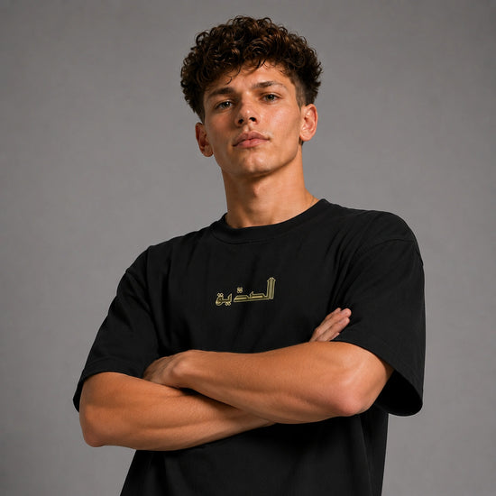 A young man with curly brown hair wearing a black crewneck t-shirt with gold Arabic calligraphy on the chest, posed with arms crossed against a solid gray studio background.