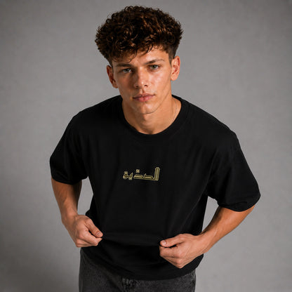 A young man with curly brown hair wearing a black short-sleeve t-shirt with gold Arabic calligraphy on the chest, posing against a neutral gray studio background.