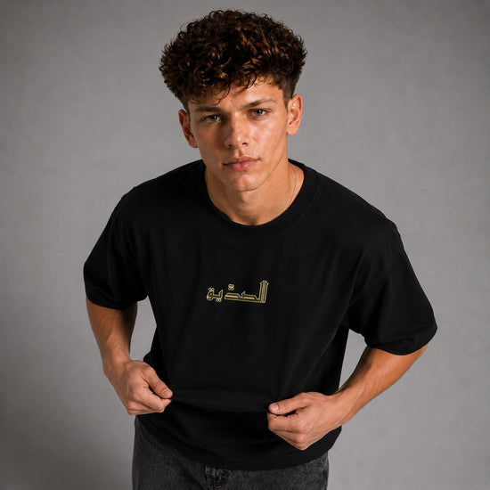 A young man with curly brown hair wearing a black short-sleeve t-shirt with gold Arabic calligraphy on the chest, posing against a neutral gray studio background.