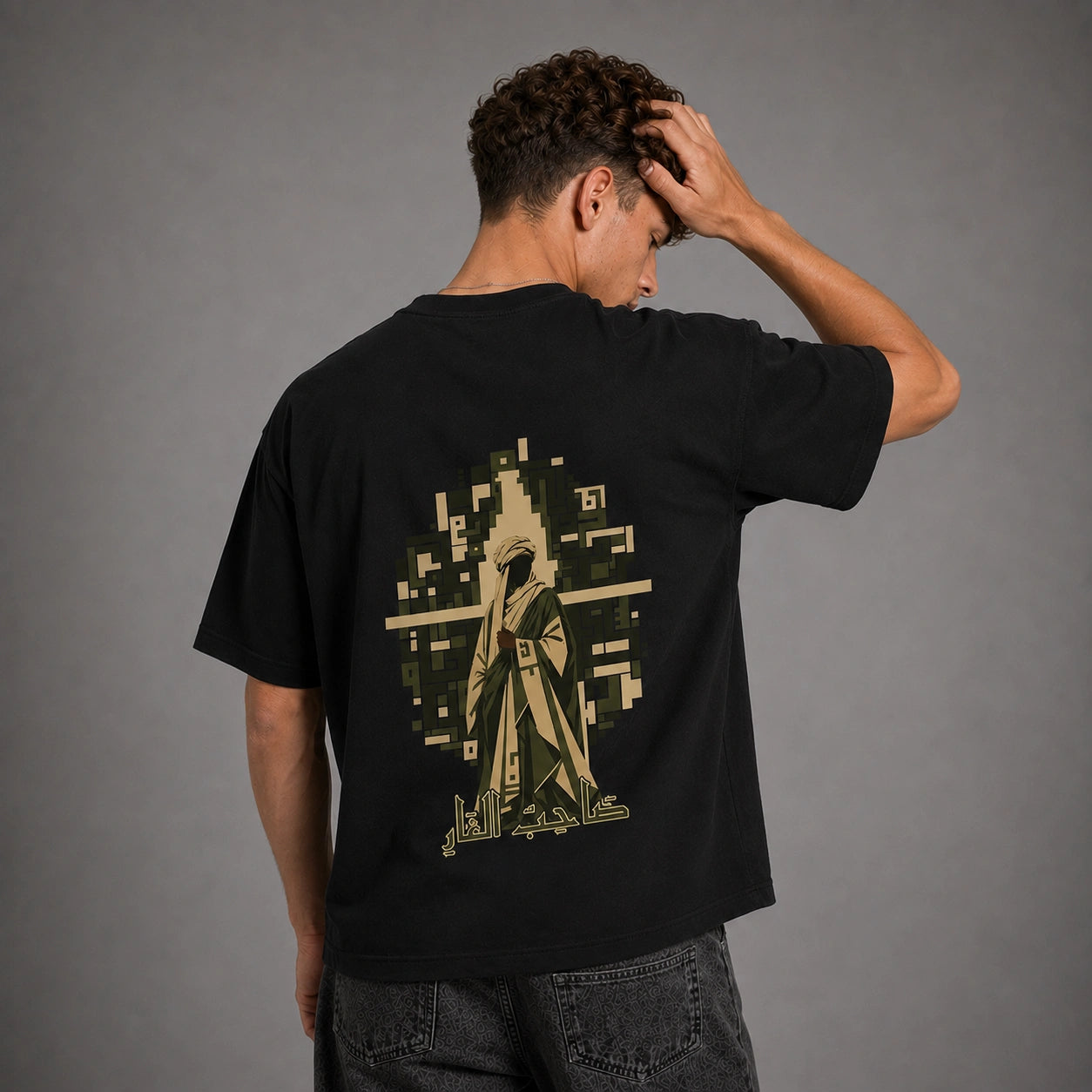 A man viewed from behind wearing a black t-shirt with a large pixelated graphic of a hooded figure in beige and olive green on the back, set against a plain gray studio background.