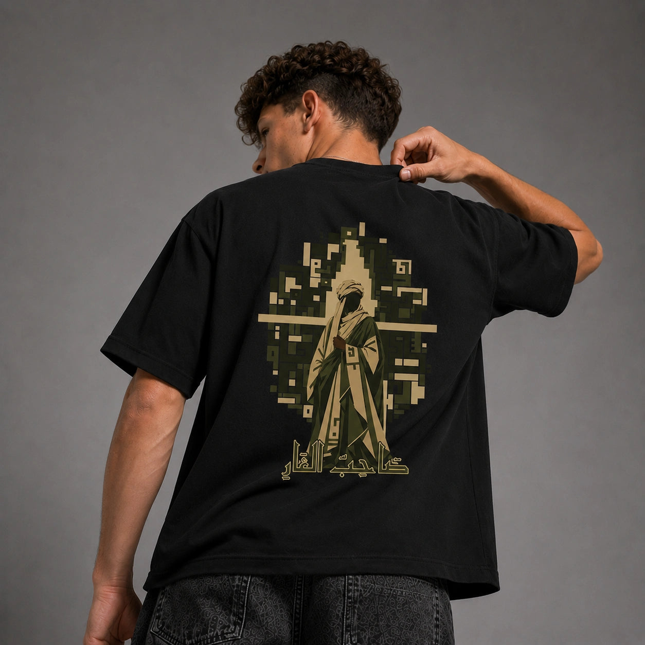A man wearing a black t-shirt from the back, showing a large graphic print of a robed figure in a pixelated geometric design in olive green and beige.