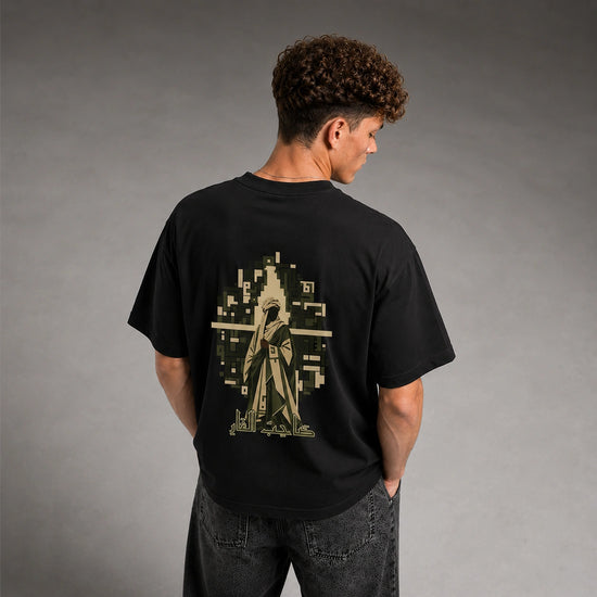 A man viewed from behind wearing a black t-shirt with a large pixelated graphic on the back in olive green and beige, paired with dark gray jeans against a gray studio background.
