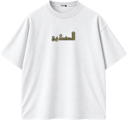 A white short-sleeve crewneck t-shirt featuring Arabic calligraphy printed in olive green and gold on the center chest.
