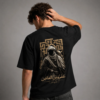 A man wearing a black t-shirt from the back, showing a large graphic print in gold and beige. The graphic features a hooded figure, a geometric pattern, and Arabic text.