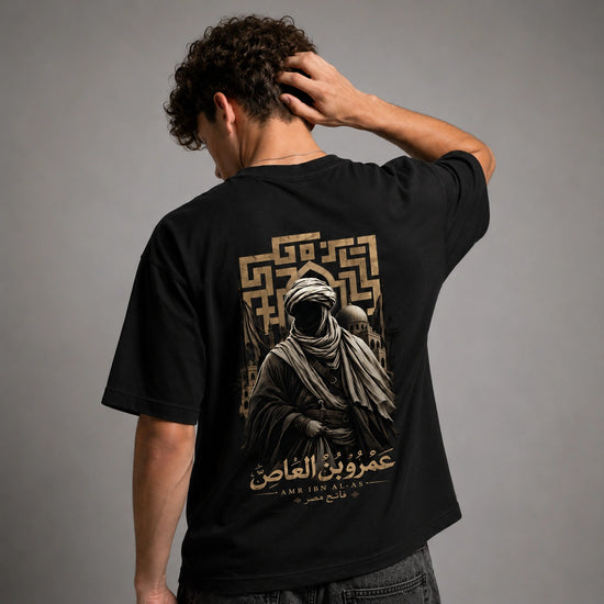 A man wearing a black t-shirt from the back, showing a large graphic print in gold and beige. The graphic features a hooded figure, a geometric pattern, and Arabic text.