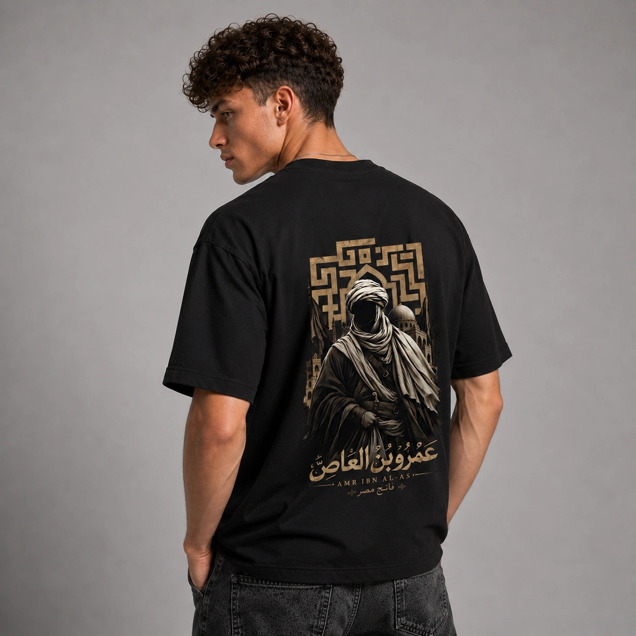 A man wearing a black t-shirt with a large graphic print on the back, photographed from behind against a neutral gray studio background.