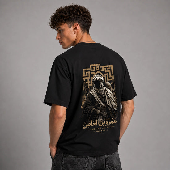 A man wearing a black t-shirt with a large graphic print on the back, photographed from behind against a neutral gray studio background.