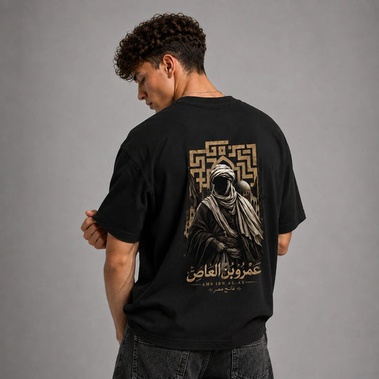 A man wearing a black graphic t-shirt, viewed from behind, showing a large gold and beige print of a hooded figure and Arabic text on the back, set against a plain gray studio background.