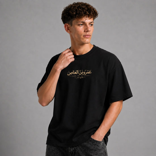 A man wearing a black short-sleeve t-shirt with gold Arabic calligraphy on the chest, posed against a solid gray studio background.