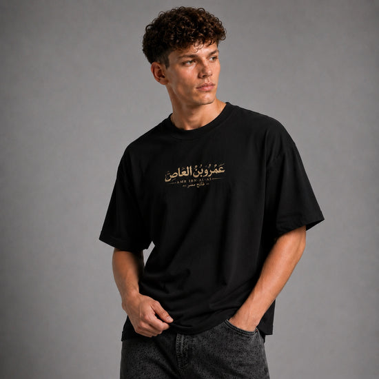 A man wearing a black short-sleeve t-shirt with gold Arabic text and 'LAW & ORDER' printed on the chest, posed against a neutral gray studio background.