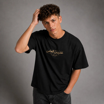 A man wearing a black short-sleeve t-shirt with gold Arabic calligraphy and English text on the chest, paired with dark gray jeans, posing against a neutral gray studio background.