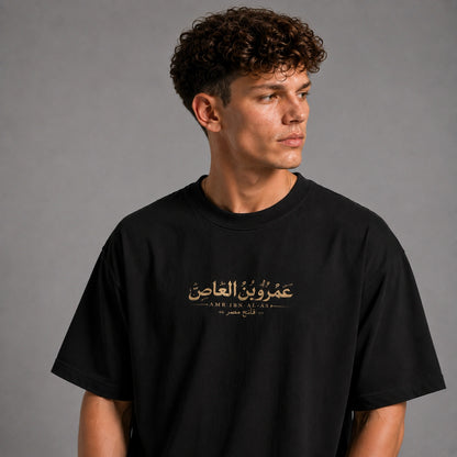 A man wearing a black crewneck t-shirt with gold Arabic text and 'AMEN ALAS' printed on the chest, photographed against a neutral gray background.