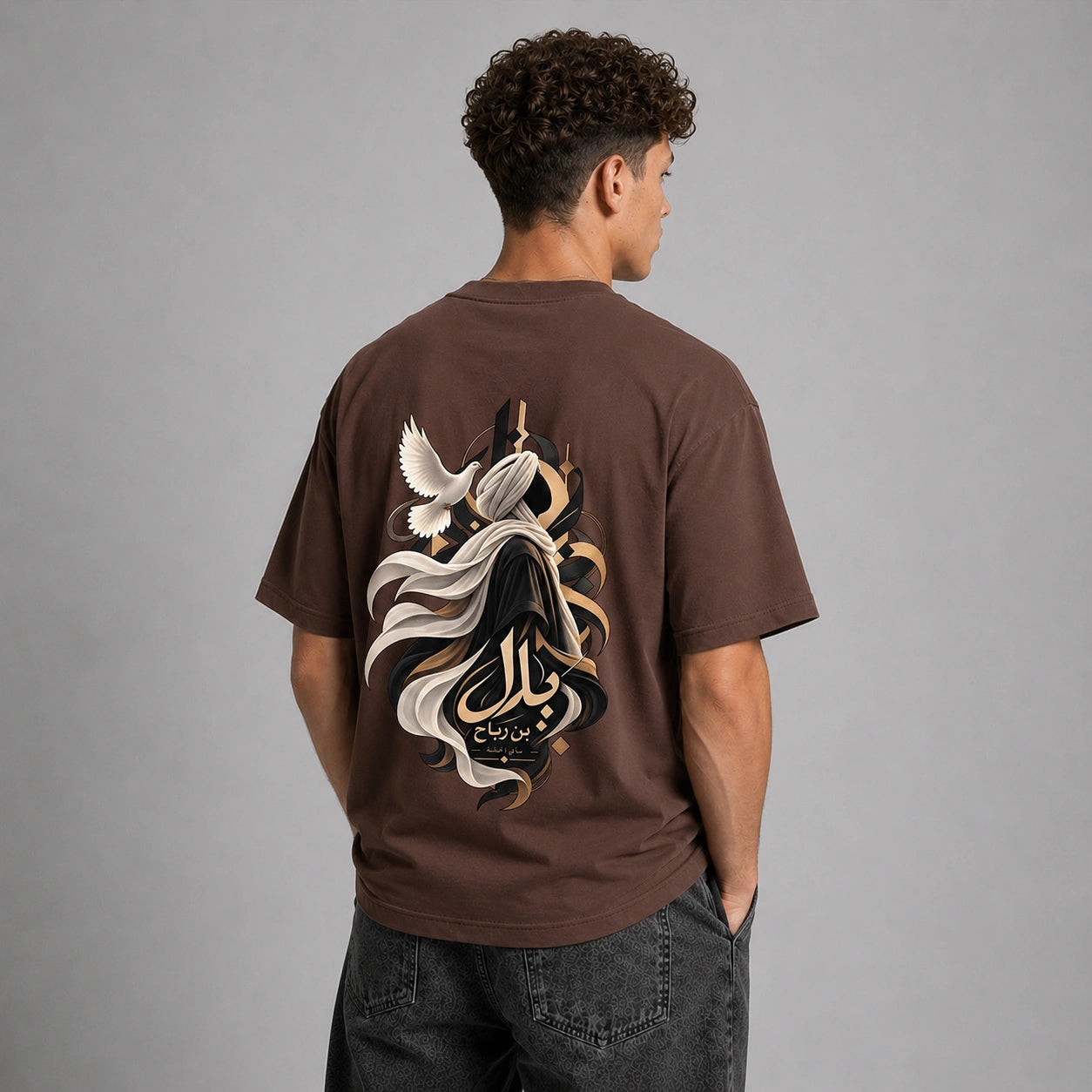 A man viewed from behind wearing a brown t-shirt with a large graphic print on the back, featuring a stylized figure, flowing white and black elements, and Arabic calligraphy. He is also wearing dark gray jeans against a solid gray studio background.