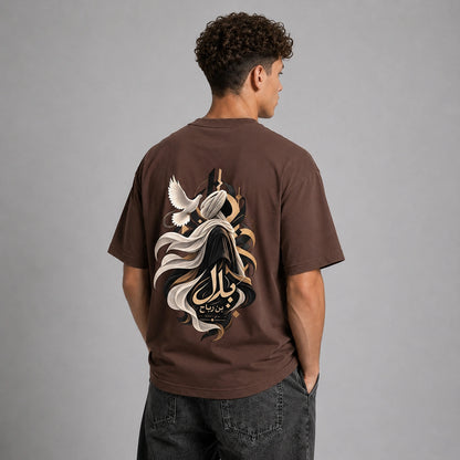 A man viewed from behind wearing a brown t-shirt with a large graphic print on the back, featuring a stylized figure, flowing white and black elements, and Arabic calligraphy. He is also wearing dark gray jeans against a solid gray studio background.
