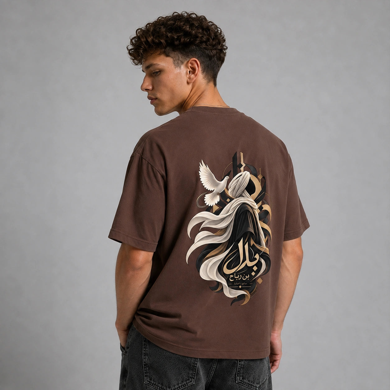 A man viewed from behind wearing a brown t-shirt with a large graphic print on the back, featuring a stylized figure, flowing white and black elements, and Arabic calligraphy. He is also wearing dark gray jeans against a solid gray studio background.