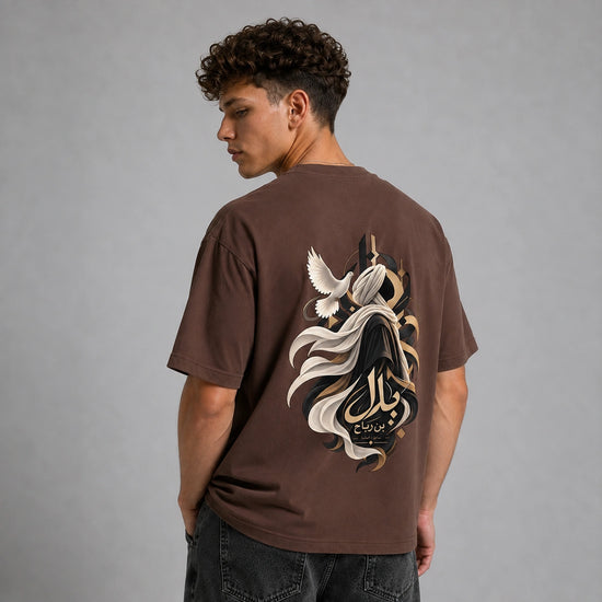 A man viewed from behind wearing a brown t-shirt with a large graphic print on the back, featuring a stylized figure, flowing white and black elements, and Arabic calligraphy. He is also wearing dark gray jeans against a solid gray studio background.