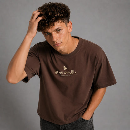 A man wearing a brown short-sleeve t-shirt with a gold Arabic text graphic on the chest, posed against a solid gray studio background.