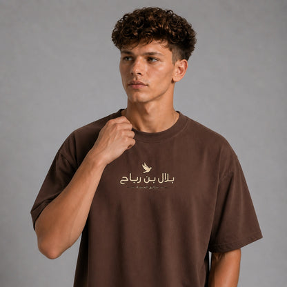 A young man with curly brown hair wearing a brown short-sleeve t-shirt with a gold Arabic text graphic on the chest, posed against a neutral gray studio background.