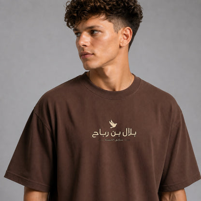 A man wearing an oversized brown t-shirt with a beige graphic print featuring Arabic text and a bird silhouette on the chest, set against a plain gray studio background.