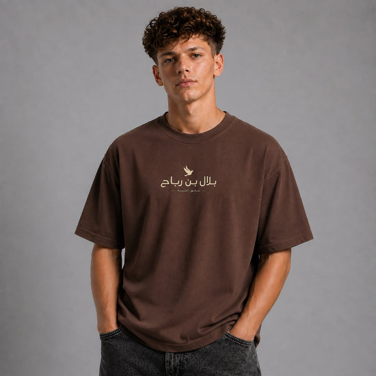 A young man with curly brown hair wearing a brown short-sleeve t-shirt with a gold Arabic graphic on the chest, paired with dark gray jeans, standing against a solid gray studio background.