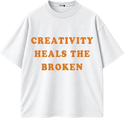 A white short-sleeve crewneck t-shirt with the phrase 'CREATIVITY HEALS THE BROKEN' printed in orange across the chest.
