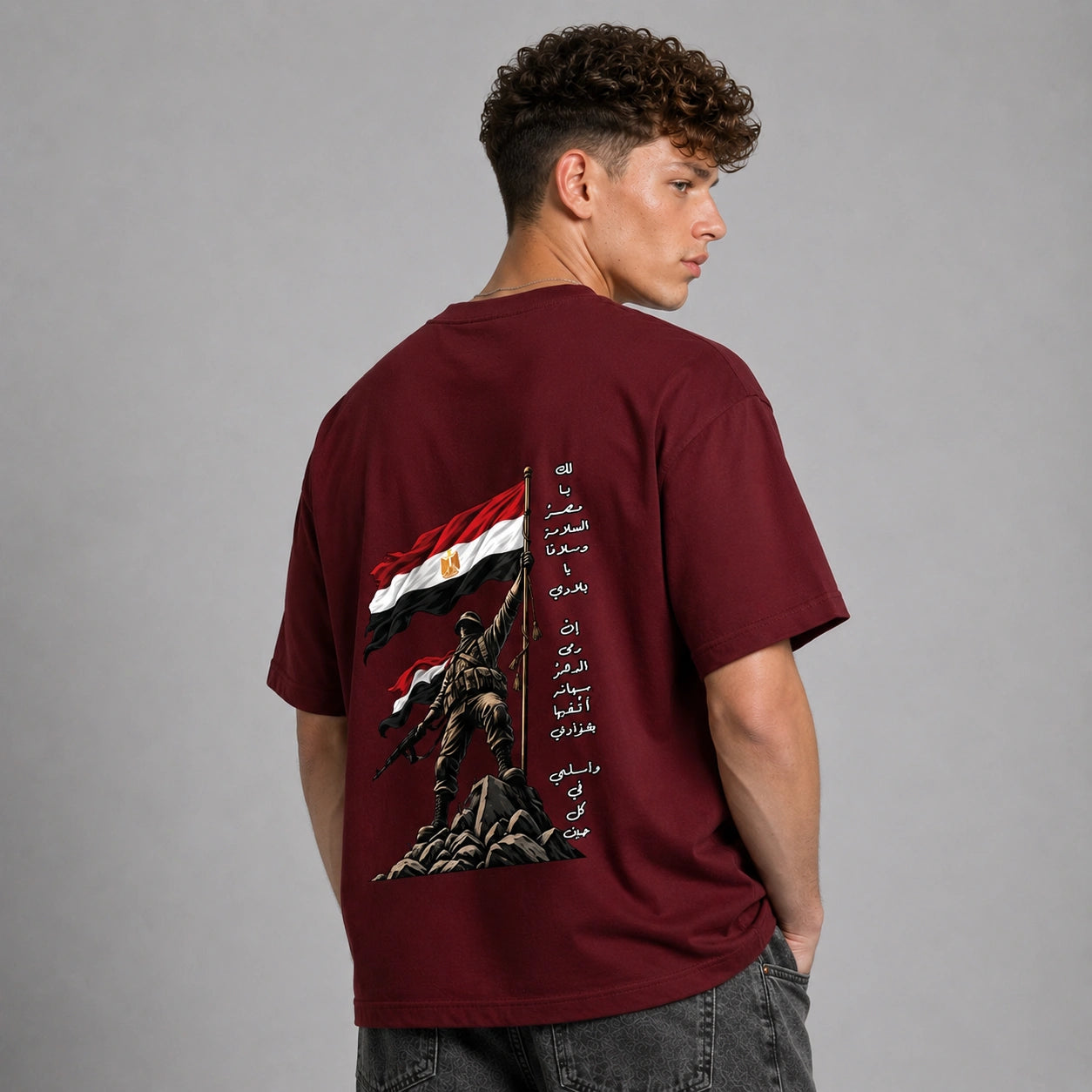 A man wearing a maroon short-sleeve t-shirt, viewed from the back. The shirt features a large graphic print of a soldier holding a flag with red, black, and white horizontal stripes and gold details, alongside vertical black Arabic text.