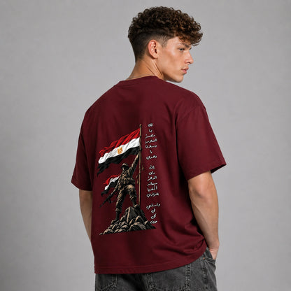 A man wearing a maroon short-sleeve t-shirt, viewed from the back. The shirt features a large graphic print of a soldier holding a flag with red, black, and white horizontal stripes and gold details, alongside vertical black Arabic text.