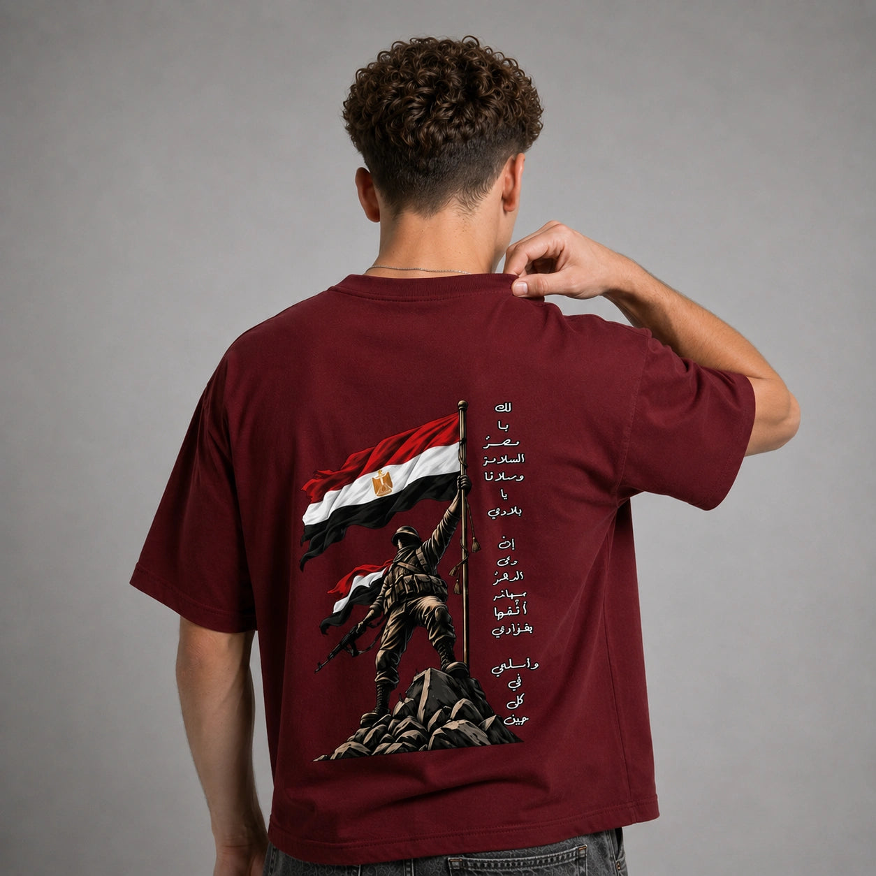 A man viewed from behind wearing a maroon short-sleeve t-shirt with a large graphic print on the back. The graphic features the Egyptian flag, a soldier holding a flagpole, and vertical Arabic text.