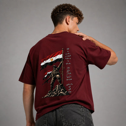 A man wearing a maroon short-sleeve t-shirt, viewed from the back. The shirt features a large graphic print of a soldier holding an Egyptian flag on a cliff, with Arabic text printed vertically on the right side.