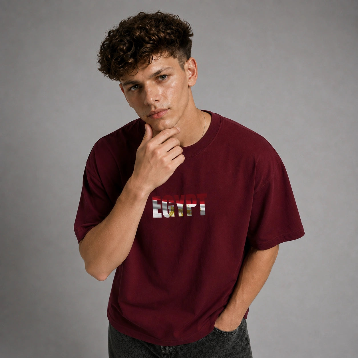 A young man with curly brown hair wearing a maroon crewneck t-shirt with a stylized 'LOVE' graphic across the chest, posed against a neutral gray studio background.
