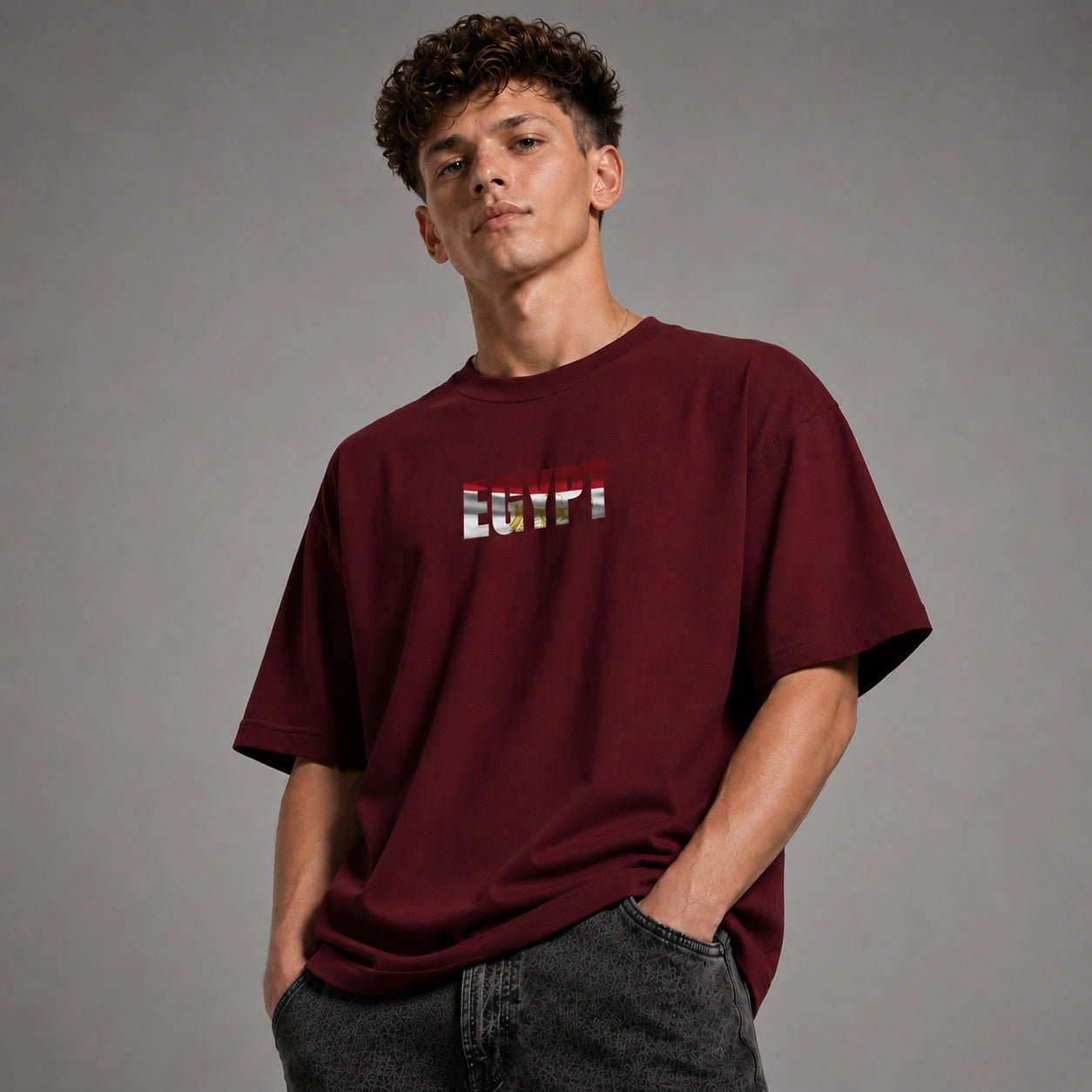 A young man wearing a burgundy t-shirt with a colorful 'BUTTI' graphic on the chest, paired with dark gray jeans, standing against a solid gray studio background.