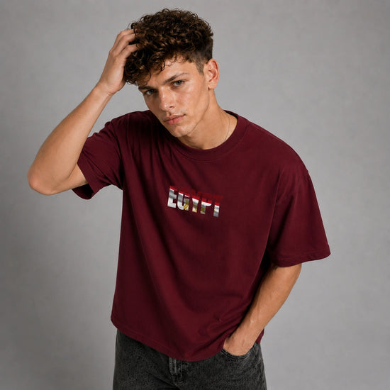 A man wearing a maroon crewneck t-shirt with a graphic on the chest, posed against a neutral gray studio background.