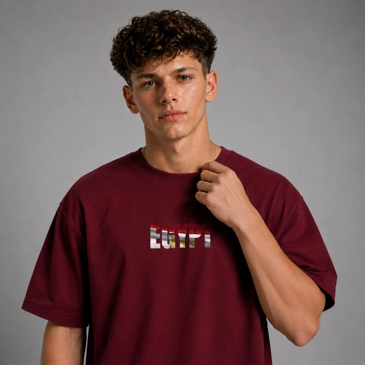 A young man with curly brown hair wearing a maroon crewneck t-shirt with a graphic text print on the chest, posing against a neutral gray background.