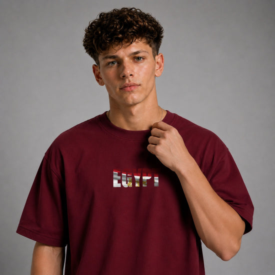 A young man with curly brown hair wearing a maroon crewneck t-shirt with a graphic text print on the chest, posing against a neutral gray background.