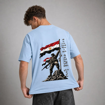 A man wearing a light blue t-shirt, viewed from the back. The shirt features a large graphic print of a soldier standing on a rock holding an Egyptian flag, with Arabic text printed vertically on the right side of the graphic.