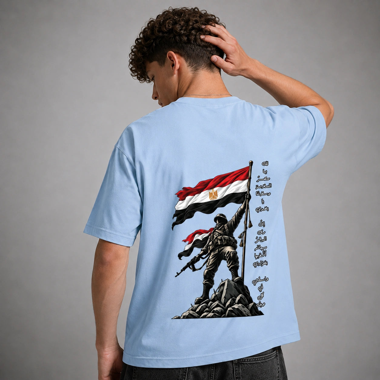 A man wearing a light blue t-shirt, viewed from the back. The shirt features a large graphic print of a soldier holding the Egyptian flag on a rocky peak, with Arabic calligraphy printed vertically on the right side of the graphic.