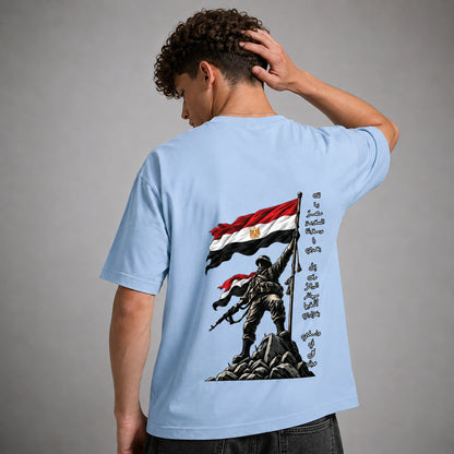A man wearing a light blue t-shirt, viewed from the back. The shirt features a large graphic print of a soldier holding the Egyptian flag on a rocky peak, with Arabic calligraphy printed vertically on the right side of the graphic.