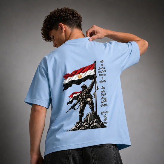 A man wearing a light blue t-shirt, viewed from the back. The shirt features a large graphic print of a soldier raising an Egyptian flag on a mountain peak, with Arabic calligraphy on the right side.