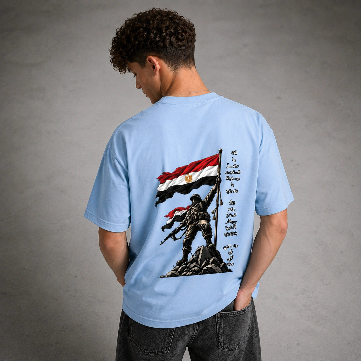 A man viewed from behind wearing a light blue t-shirt with a large graphic print on the back. The graphic features a soldier standing on rocks holding an Egyptian flag, with Arabic calligraphy on the right side.