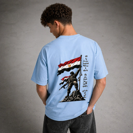 A man viewed from behind wearing a light blue t-shirt with a large graphic print on the back. The graphic features a soldier standing on rocks holding an Egyptian flag, with Arabic calligraphy on the right side.