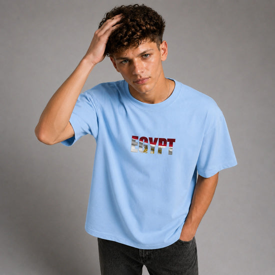 A young man wearing a light blue t-shirt with 'EGYPT' printed across the chest in a stylized, colorful font, posed against a neutral gray studio background.
